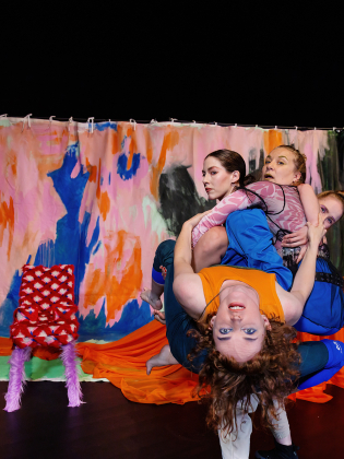 Foto: Petra Älvstrand Fyra kvinnor håller i varandra framför en svart scen med en färgglad fond och ett bord med orange duk.