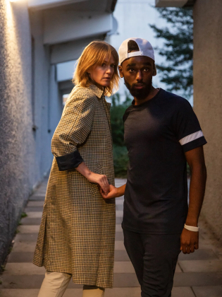 Kvinna i lång kappa och man i mörk t-shirt och keps håller varandra i handen i en smal gränd.