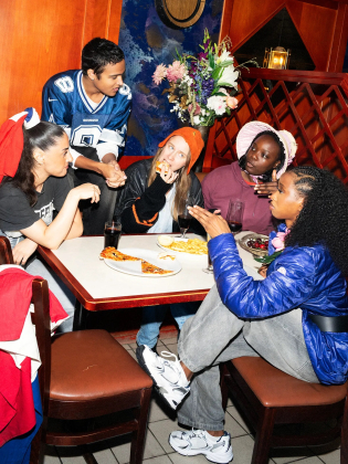 Fem unga personer sitter vid ett bord med mat, pratar och äter i en restaurangmiljö.