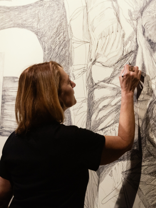 Kvinna med axellångt brunt hår tecknar med kol på en stor vägg täckt av detaljerade skisser.