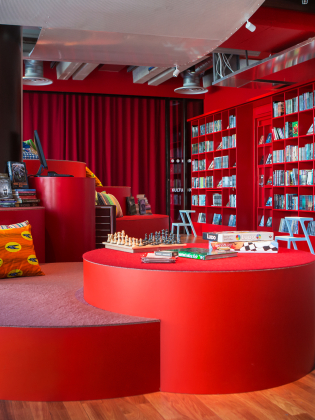 Modernt bibliotek med röda bokhyllor, rund sittplats med kuddar, sällskapsspel och schackbräde på bordet.