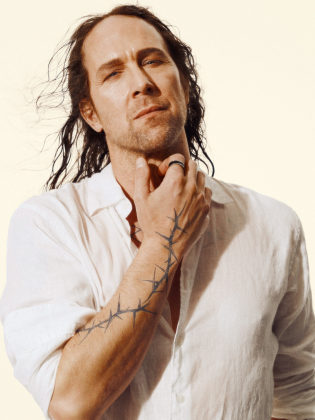 Man i 40-årsåldern med långt mörkt hår, vit skjorta och taggtrådsliknande tatuering på höger arm.