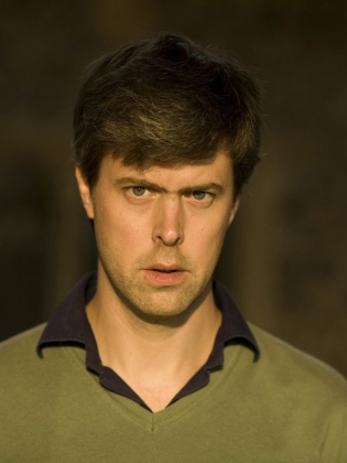 Man i 30–40-årsåldern med kort brunt hår i grön tröja med blå krage mot mörk bakgrund.