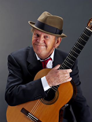 Mikael Neumann leendes iklädd kostym och hatt med en gitarr i handen.