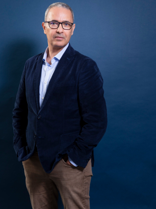 Man i 50–60-årsåldern med kort grått hår och glasögon, i blå kavaj mot blå bakgrund.