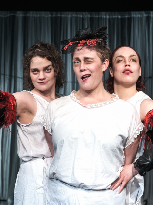 Tre kvinnor i vita underkläder, två med svarta ärmar med röda fjädrar, poserar framför ett draperi på en scen.