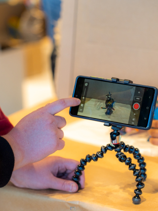 Två barnhänder filmar lerafigur med mobilkamera monterad på stativ.