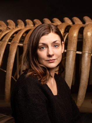 Kvinna i 30-årsåldern med brunt axellångt hår och svart tröja, står framför valskelett.