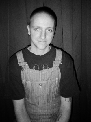 Ung person med rakat huvud, örhängen och jeanshängselbyxor poserar mot mörk bakgrund.