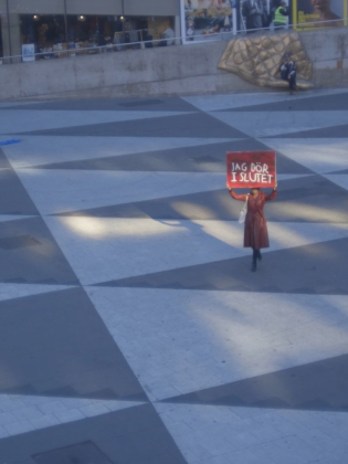 Person i röd kappa står ensam på torg och håller skylt med texten "JAG DÖR I SLUTET".