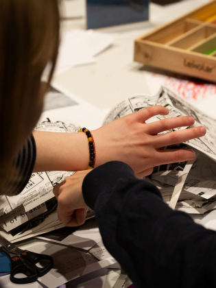 Två händer bläddrar i serietidningar vid ett bord med färgpennor, sax, stämpel och foton.