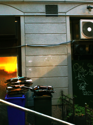 Två överfulla papperskorgar med buntar av papper står vid en husvägg med ett fönster som reflekterar en orange solnedgång.