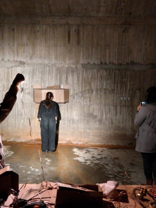 Tre personer i mörkt rum, en kvinna står mot vägg med kartong över huvudet, två andra dokumenterar med teknik.