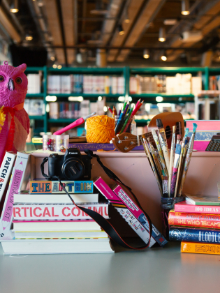 Bord fullt av färgglada konstmaterial, böcker, penslar, kamera och en rosa uggla i ett bibliotek.