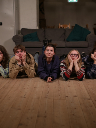 Fem unga personer ligger på golvet med hakan i händerna framför en soffa i ett vardagsrum.