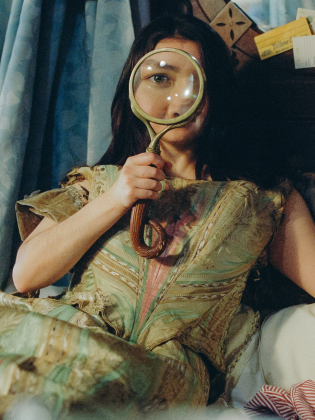 Kvinna ca 30–40 år med långt mörkt hår i grön vintageklänning håller förstoringsglas framför ena ögat i säng.