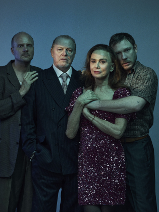 Fyra vuxna står tätt: skallig man i mörk kavaj, äldre man i kostym, kvinna 50–60 i röd paljettklänning, ung man håller om henne.