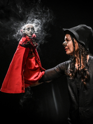 Vuxen kvinna i hatt och glasögon håller en liten docka i röd mantel på en mörk teaterscen.