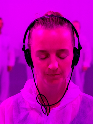 Kvinna i 30-årsåldern med ljust uppsatt hår och hörlurar, vit hoodie, blundar i rosa ljus, suddiga personer i vitt bakom.