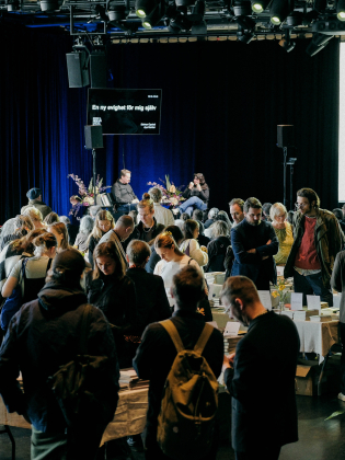 Många vuxna besökare bläddrar bland böcker på bord; en man och en kvinna sitter på scen i bakgrunden.