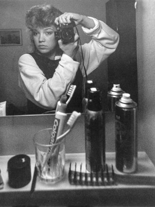 Ung kvinna med kort lockigt hår tar spegelselfie med kamera i badrum; tandborste, tandkräm och sprayburkar på handfat.