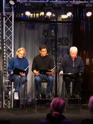 Sex personer, tre män och tre kvinnor i olika åldrar, sitter på pallar på scen och läser ur svarta manus.