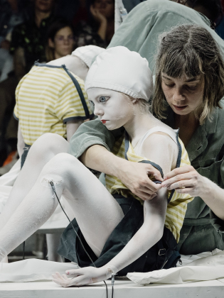 Kvinnlig tekniker i grön rock justerar vitmålad flicka i randig tröja med kablar, publik i bakgrunden.