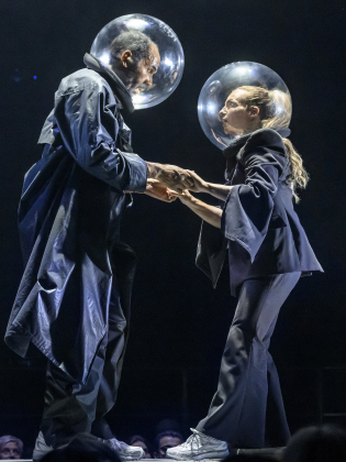 Man och kvinna i mörka kläder och stora genomskinliga hjälmar håller varandras händer på scen.