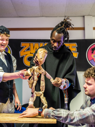 Tre personer samarbetar kring en docka av papper i ett rum, de skrattar och fokuserar under en workshop eller repetition.