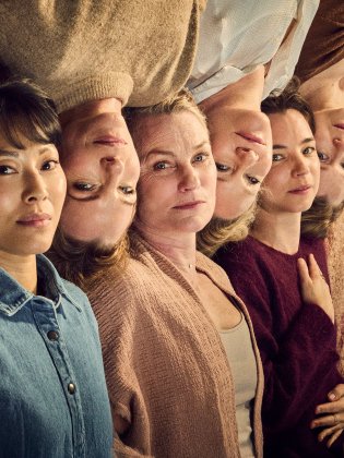 Sex kvinnor ligger tätt sida vid sida med huvuden mot varandra, sedda uppifrån mot en varmtonad bakgrund; de har olika åldrar och uttryck och blickar lugnt mot kameran.