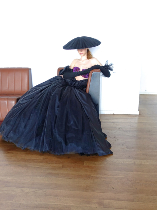 Person i svart, voluminös klänning och bred hatt sitter i fåtölj i ett ljust rum med trägolv och vita väggar.