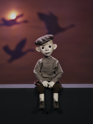 Docklik figur med basker sitter på en kant i dämpat ljus, med skuggor av flygande figurer projicerade på väggen bakom.