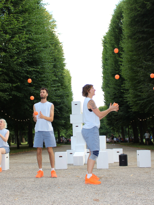 Tre personer i ljusa kläder jonglerar med orange bollar på en grusgång mellan höga träd.