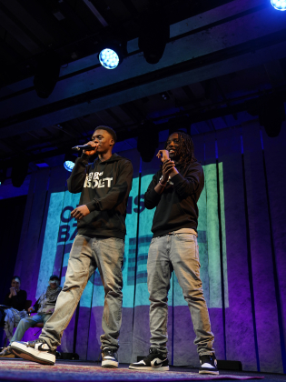 Två personer står på scen med mikrofoner och uppträder under blå scenbelysning inför publik.