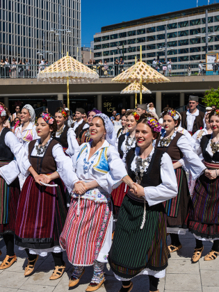 Dansare klädda i serbiska folkdräkter som dansar tillsammans på ett soligt torg.