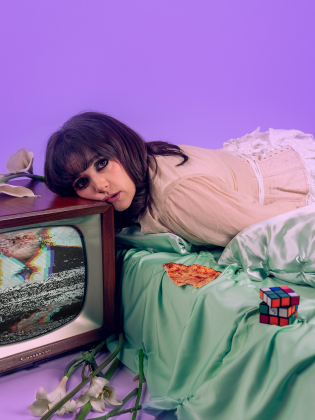 Kvinna ligger på säng med huvudet mot en tv med brus; pizza, Rubiks kub och blommor runtom i scen med lila bakgrund.