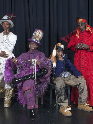 Fyra personer poserar i färgstarka, extravaganta outfits och hattar framför en mörk scenridå.