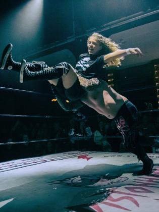 Två personer brottas i en wrestling ring upplysta av strålkastare. Den ena personen kastas genom luften medan den andra håller greppet.