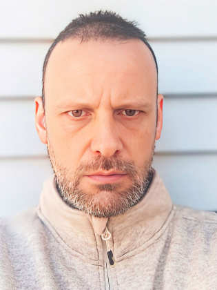 Närbild av en man med kortklippt hår och skäggstubb som ser rakt in i kameran med ett allvarligt uttryck. Han bär en ljus, högkragad tröja och står framför en enkel, ljus bakgrund med horisontella linjer.