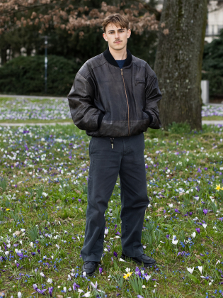 Nelio Biedermann står i ett blommande grönområde i ett avslappnat helkroppsporträtt som kombinerar ungdomlig energi med stillhet.