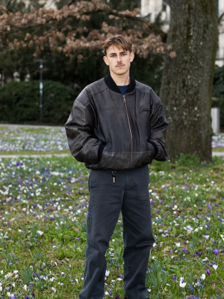 Nelio Biedermann står i ett blommande grönområde i ett avslappnat helkroppsporträtt som kombinerar ungdomlig energi med stillhet.