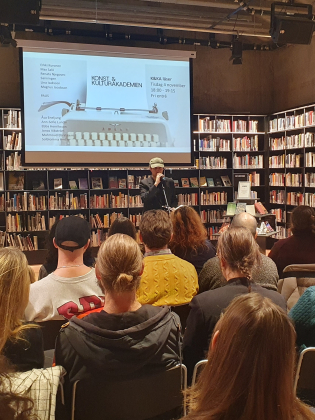 Publik sitter framför en scen i ett bibliotek. På scenen står en person med mikrofon och bakom visas en presentation på en duk.
