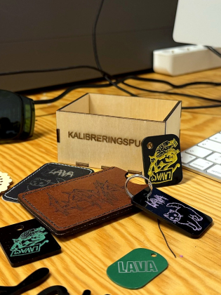 Laserskurna och graverade föremål i trä, läder och plast ligger på ett arbetsbord bredvid ett tangentbord.