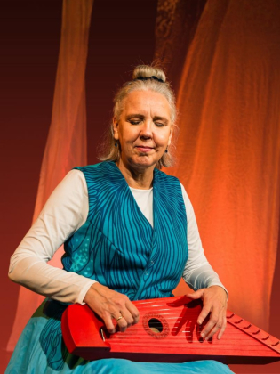Kvinna i turkos klänning sitter på en scen och spelar ett rött stränginstrument, med orange tygstycken i bakgrunden.