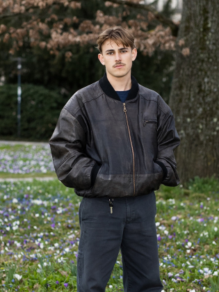 Nelio Biedermann står i ett blommande grönområde i ett avslappnat helkroppsporträtt som kombinerar ungdomlig energi med stillhet.