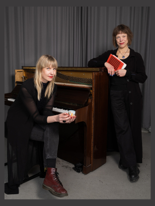 Två kvinnor klädda i svart vid ett öppnat piano. Den ena sitter framför pianot och den andra står bredvid med en bok i handen.