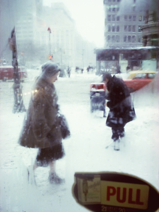 Två personer går genom ett snöigt stadslandskap i kraftigt snöfall, fotograferade genom en glasdörr med skylten ”PULL”.