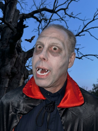 En man utklädd till Dracula med vampyrtänder, blekt ansikte, mörka ögon och svart mantel med röd krage. I bakgrunden syns ett kalt träd och en fladdermus mot en blå himmel.