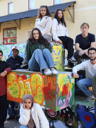 Elva ungdomar poserar tillsammans på och omkring färgglada graffitimålade betongblock framför en gul byggnad.