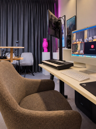 Vy från kontrollplatsen i en podcaststudio med dator, bildskärmar, keyboard och kamerautrustning. I bakgrunden syns två fåtöljer med mikrofoner vid ett runt bord.